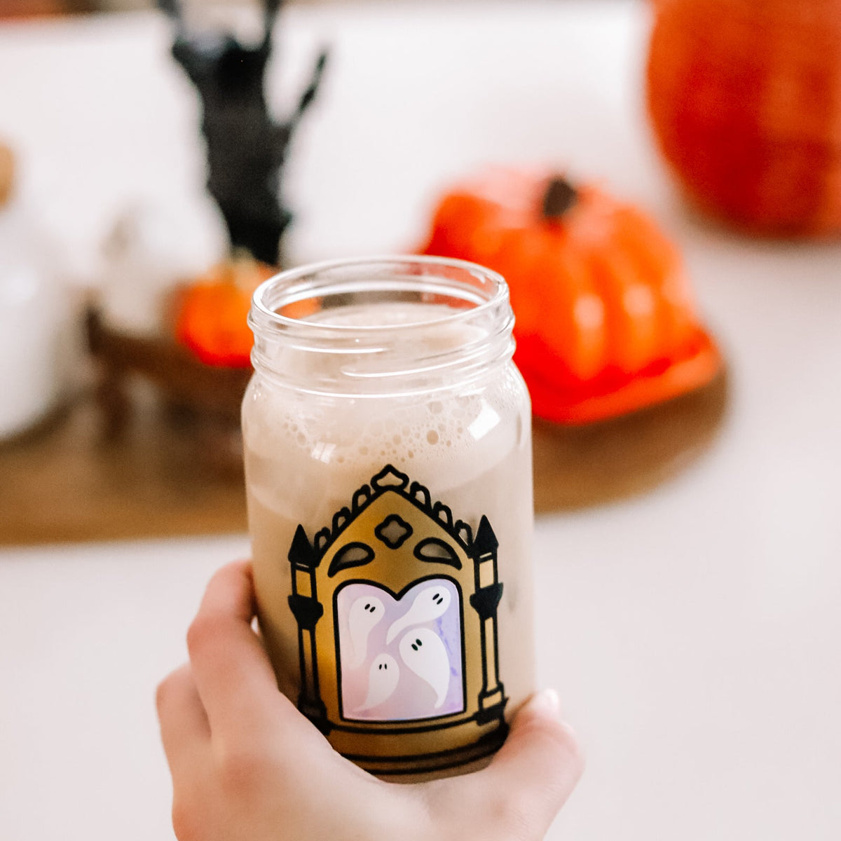 A hand holding a clear glass mason jar filled with an iced coffee with a black and gold gothic mirror design featuring white ghosts inside. The background is a softly blurred Halloween-themed table setting with pumpkins and a black skeleton hand decoration.