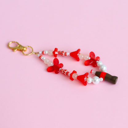 Keychain with assorted red and white beads, including red butterflies and a cola bottle, against a pink background