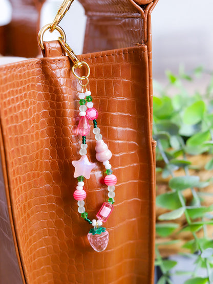 Beaded keychain with pink and green assorted beads and a light pink strawberry pendant hooked on a brown leather purse