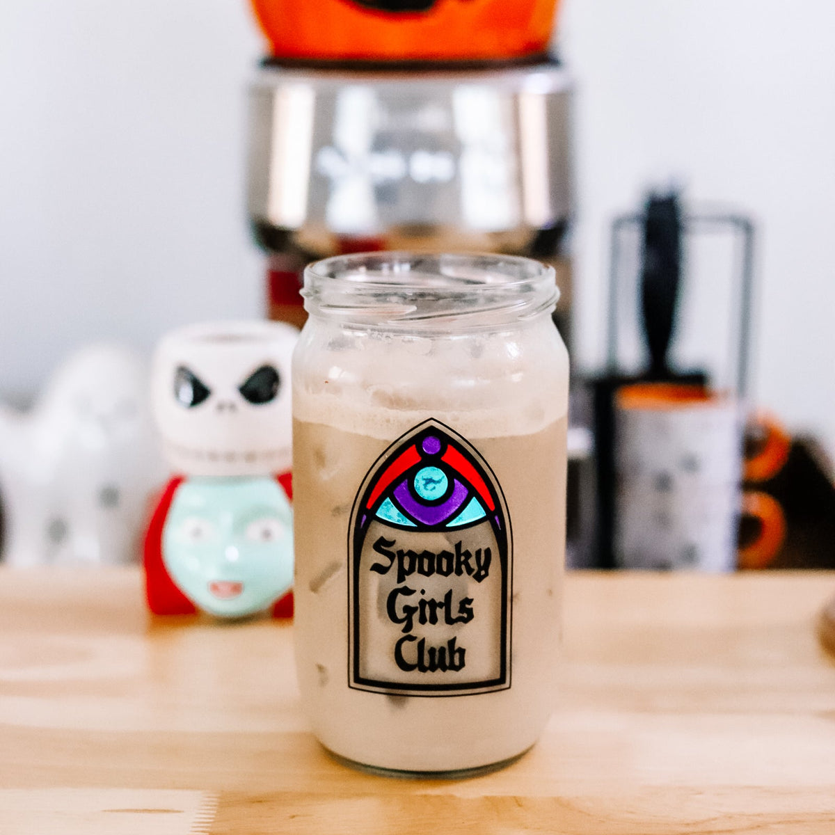A clear glass filled with an iced coffee sits on a wooden counter. The jar has a decorative gothic window design that reads 'Spooky Girls Club.'