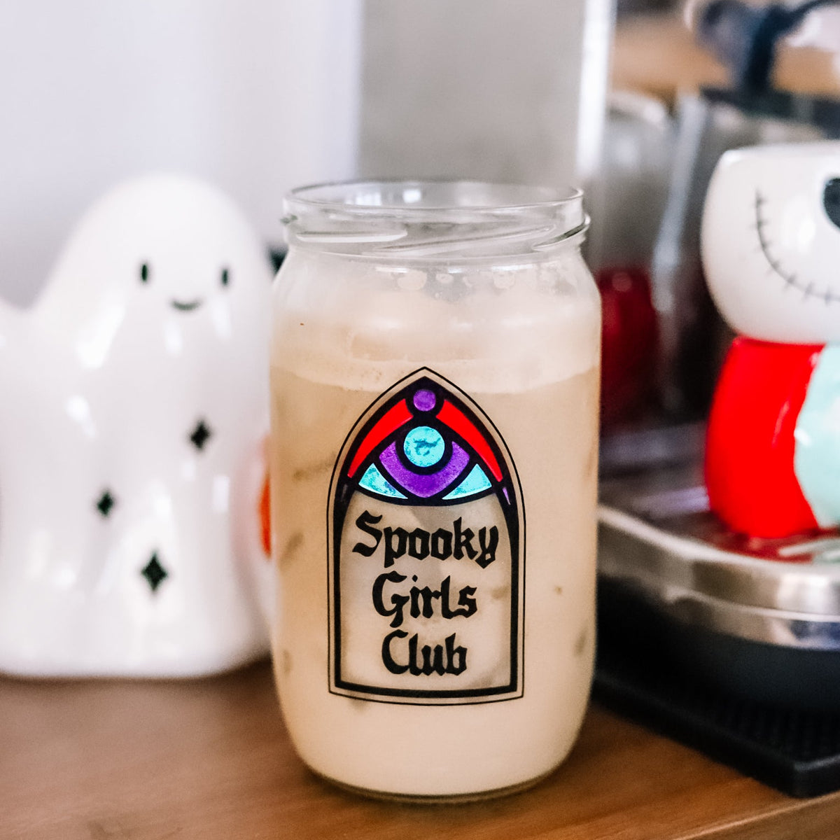 A clear glass filled with an iced coffee, featuring a gothic arch-shaped design that reads 'Spooky Girls Club' in black lettering. The jar is set on a wooden table with a white ghost-shaped ceramic object on the left and a coffee machine in the background.