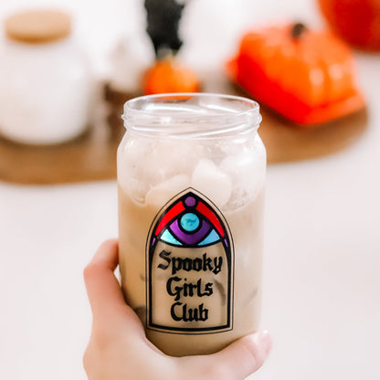 A hand holds a clear glass jar filled with an iced coffee. The jar has a decorative arch stained-glass-style design that reads 'Spooky Girls Club.'