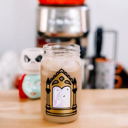 A clear glass jar filled with an iced coffee featuring a decorative gold and black gothic mirror design with white ghosts inside the mirror. The jar is set on a wooden table with a blurred Halloween-themed background.