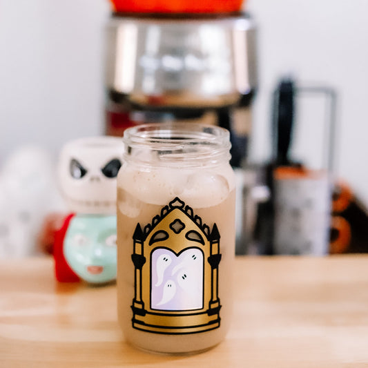 A clear glass jar filled with an iced coffee featuring a decorative gold and black gothic mirror design with white ghosts inside the mirror. The jar is set on a wooden table with a blurred Halloween-themed background.